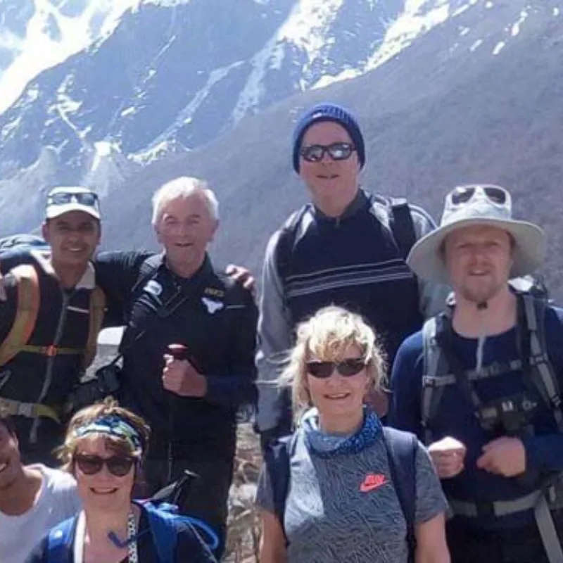 A group of five smiling trekkers, wearing sun hats and sunglasses, pose together on a rocky mountain trail with a snowy range in the background.