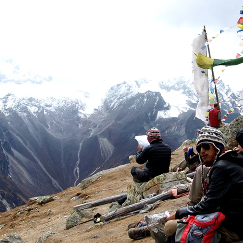 A trekker with a patterned beanie consults a map while resting on a high mountain pass with colorful prayer flags and snowy peaks in the background.