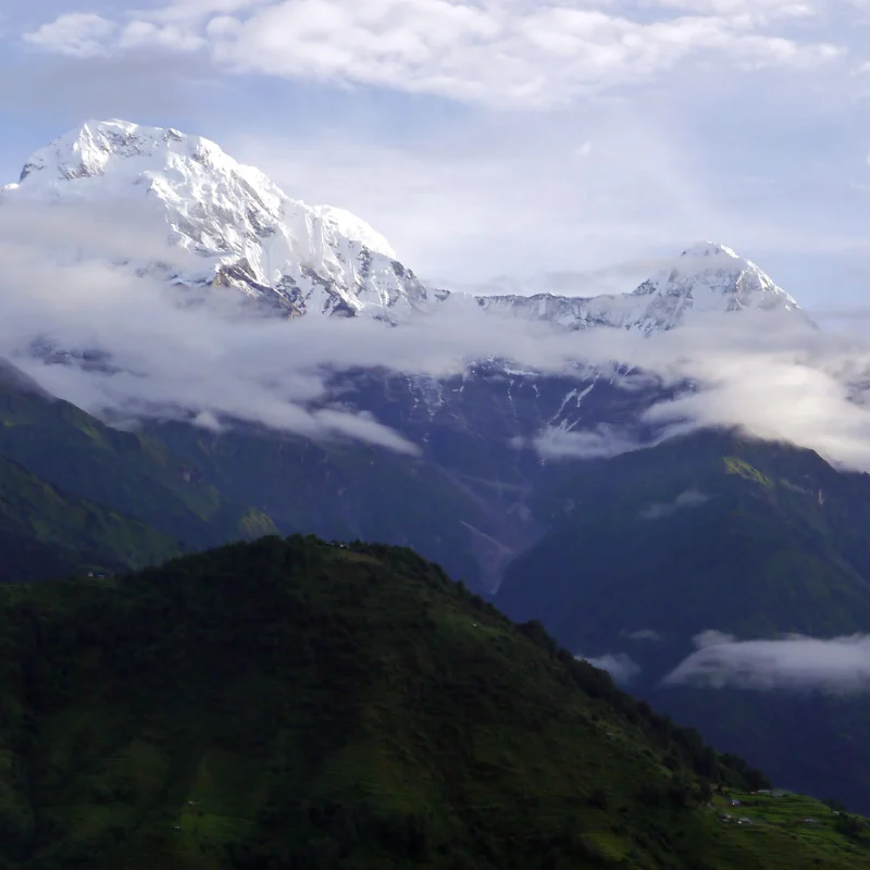 Towering snow-capped mountain peaks rising above deep green foothills, partially shrouded in low-hanging white clouds.