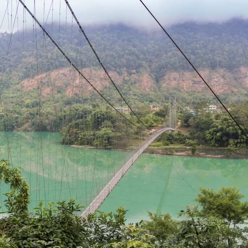 Long suspension footbridge stretching across a turquoise river surrounded by lush green forested hills in Nepal.
