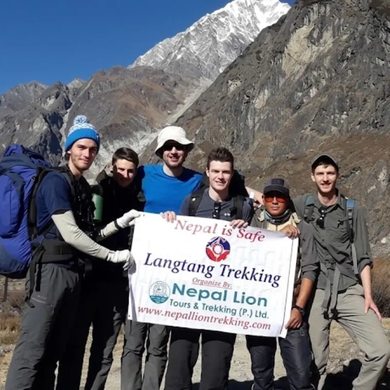 Trekkers and their local guide standing outdoors on a mountain trail holding a "Nepal is Safe Langtang Trekking" banner beneath towering Himalayan peaks.