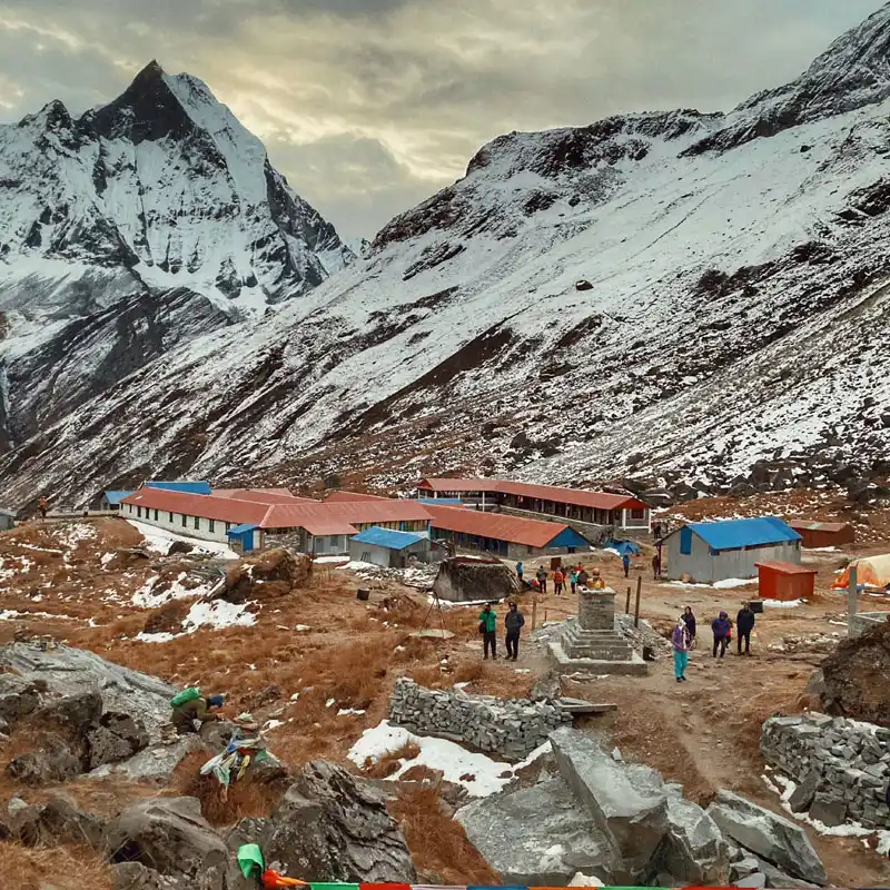The iconic Annapurna Base Camp welcome sign at 4130 meters, surrounded by colorful Buddhist prayer flags and the snow-capped peaks of the Himalayas in Nepal.