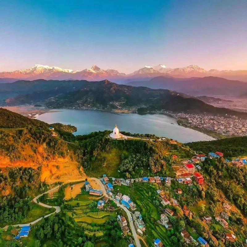 Panoramic aerial view of the Pokhara valley featuring Phewa Lake and the iconic white Shanti Stupa Peace Pagoda perched on a green forested hill, set against a stunning backdrop of the snow-capped Annapurna mountain range at sunset, Nepal.