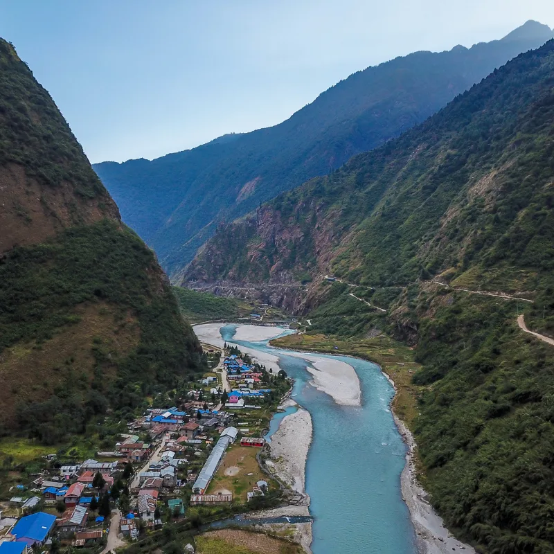 Aerial view of the brightly roofed Tal Village nestled beside a winding turquoise river deep within a steep, green Himalayan mountain valley.