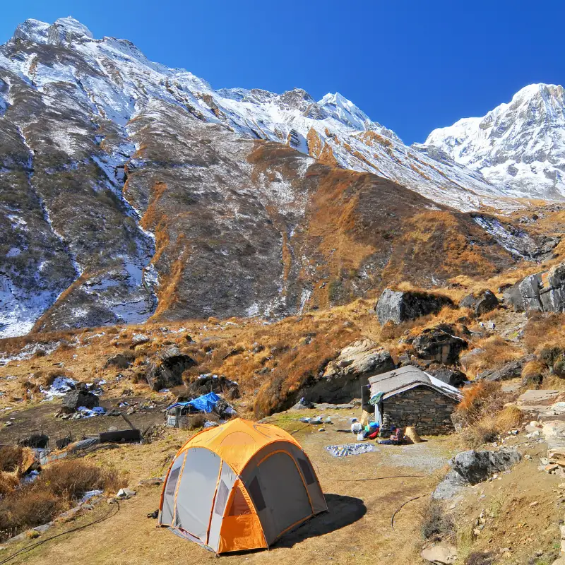 Orange camping tent and stone lodge settlement at Machhapuchhre Base Camp surrounded by snow-covered Himalayan peaks.