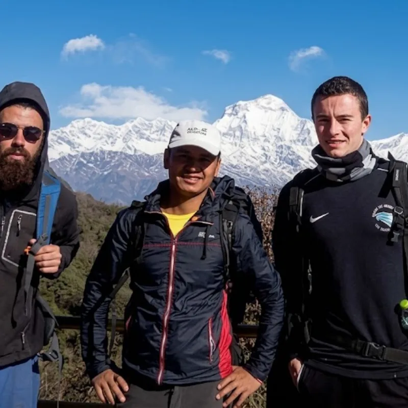 Three hikers with backpacks standing before the snow-covered Himalayan mountain range on a clear sunny day.