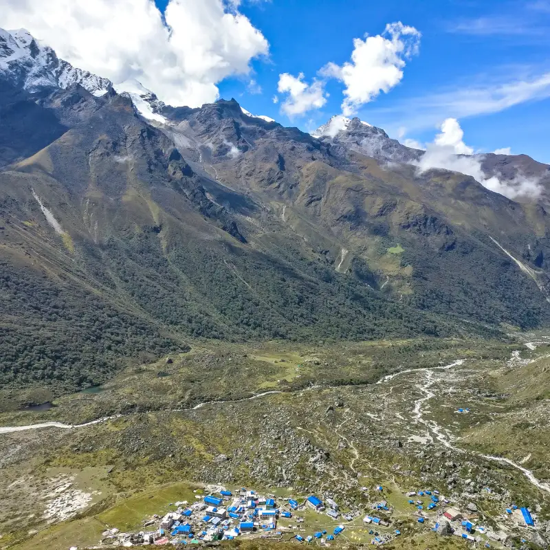Aerial view of a remote Himalayan settlement with blue-roofed buildings nestled in a vast green valley below steep rocky mountain peaks.