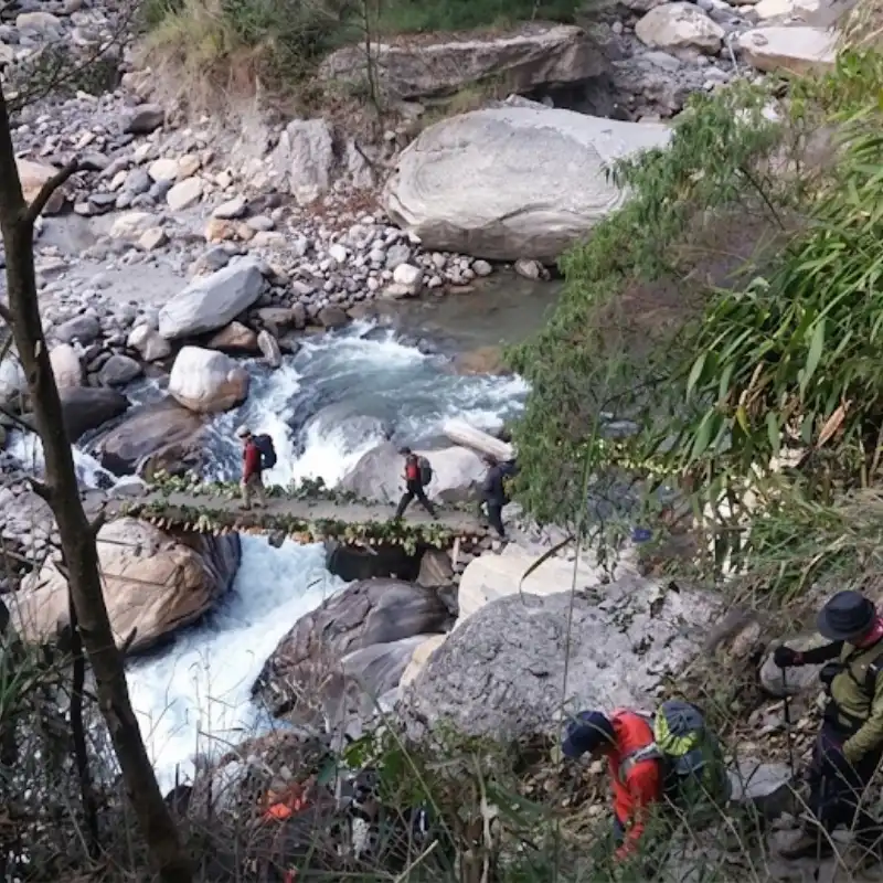 Trekkers with backpacks carefully crossing a narrow wooden footbridge over a rushing mountain river with large boulders.