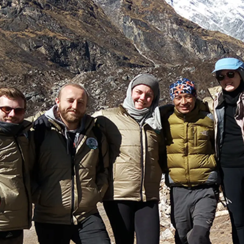 Four trekkers, including a bearded man and a woman in a white headscarf, smile together against a steep, rugged mountain slope.