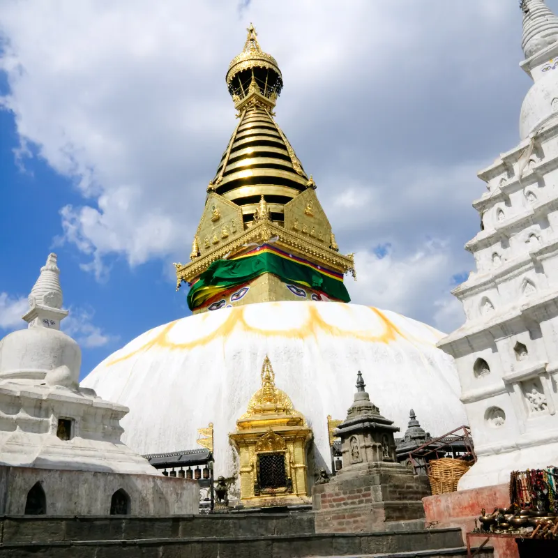 Detailed close-up perspective shot of the elaborate golden spire of the Swayambhunath Stupa, also known as the Monkey Temple, decorated with colorful prayer flags and the iconic "Buddha Eyes" symbol, against a partly cloudy blue sky in Kathmandu, Nepal.