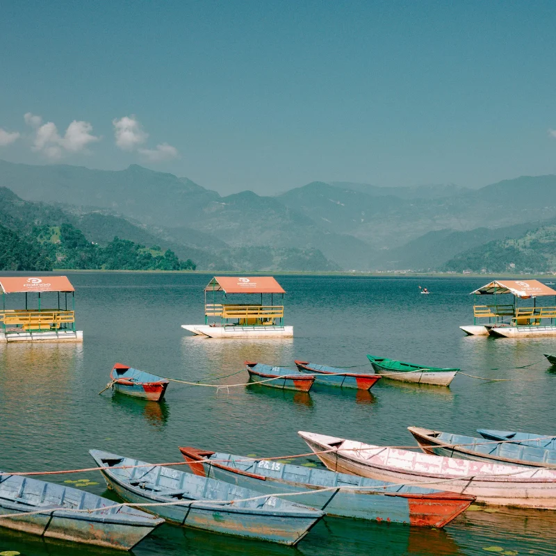 Colorful wooden boats floating on the calm blue waters of Phewa Lake in Pokhara against a backdrop of hazy hills.