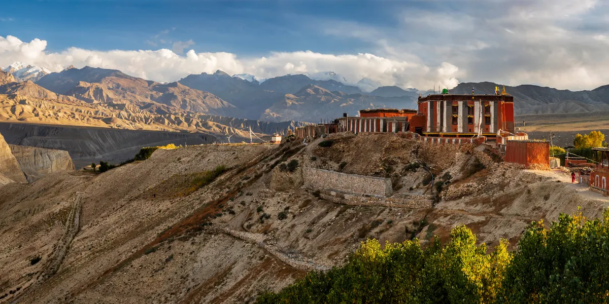 Traditional red Tibetan Buddhist monastery perched on a high ridge overlooking arid hills and distant snow-capped mountains.
