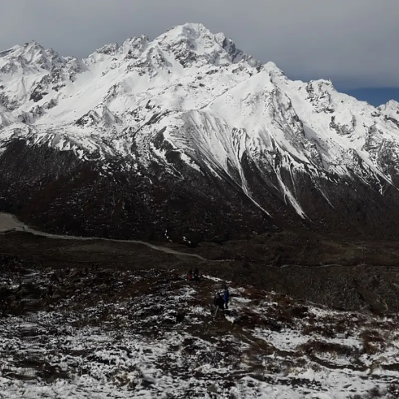 Snow-covered mountain peak with steep ridges and trekkers traversing an icy trail in the foreground.